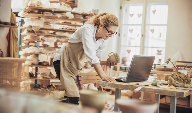 A small business owner of a pottery studio is holding a piece they made in one hand and using the other to check on an online order on their laptop in their studio.