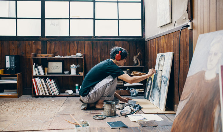 A young man is listening to a podcast while wearing large red over the ear headphones as he works on a painting in his studio.