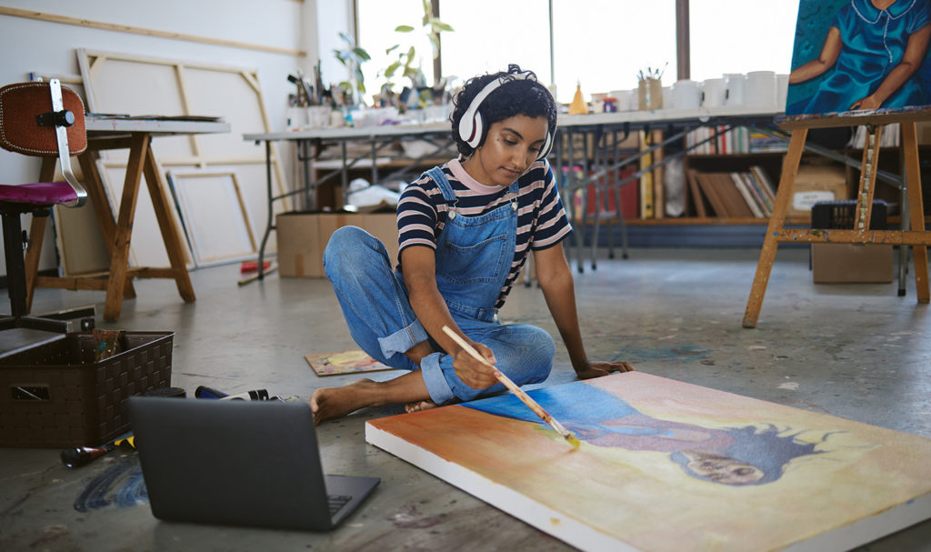 A young artist is sitting on the floor of her studio working on a painting as she listening to a podcast with her over the ear headphones on that are streaming from her laptop next to her.