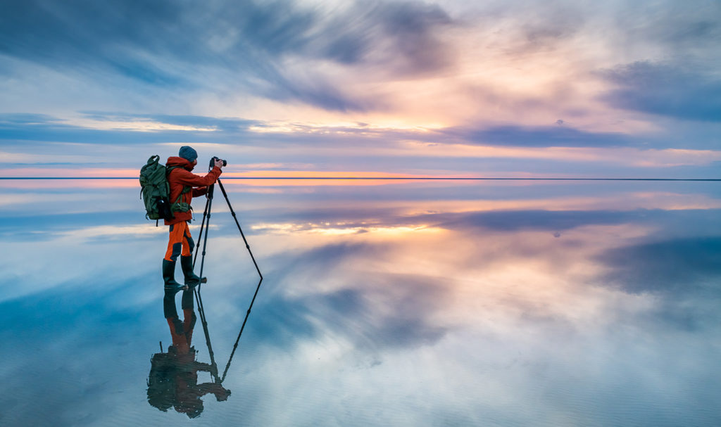 A wide shot of a nature photographer capturing a landscape photo of a cloudy sunset on a cold and empty beach. The photos he later prints and sells can be insured with ACT photography insurance.