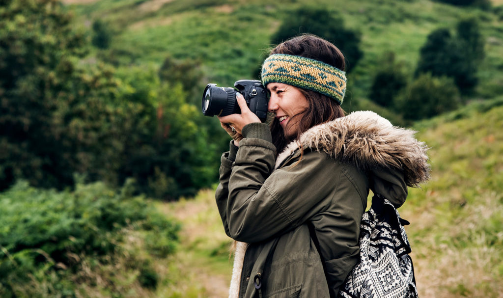 The side profile view of a young female photographer taking photos on her camera outdoors amongst green hills. Her photographers insurance can cover the photos she takes to later print and sell.