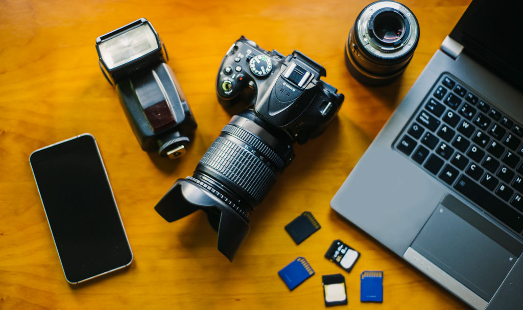 An overhead shot of a camera, smartphone, flash attachment, an extra camera lens, and memory cards are sitting on a wood desk next to a laptop. Photography insurance may be able to insure this type of equipment used in a photo business.