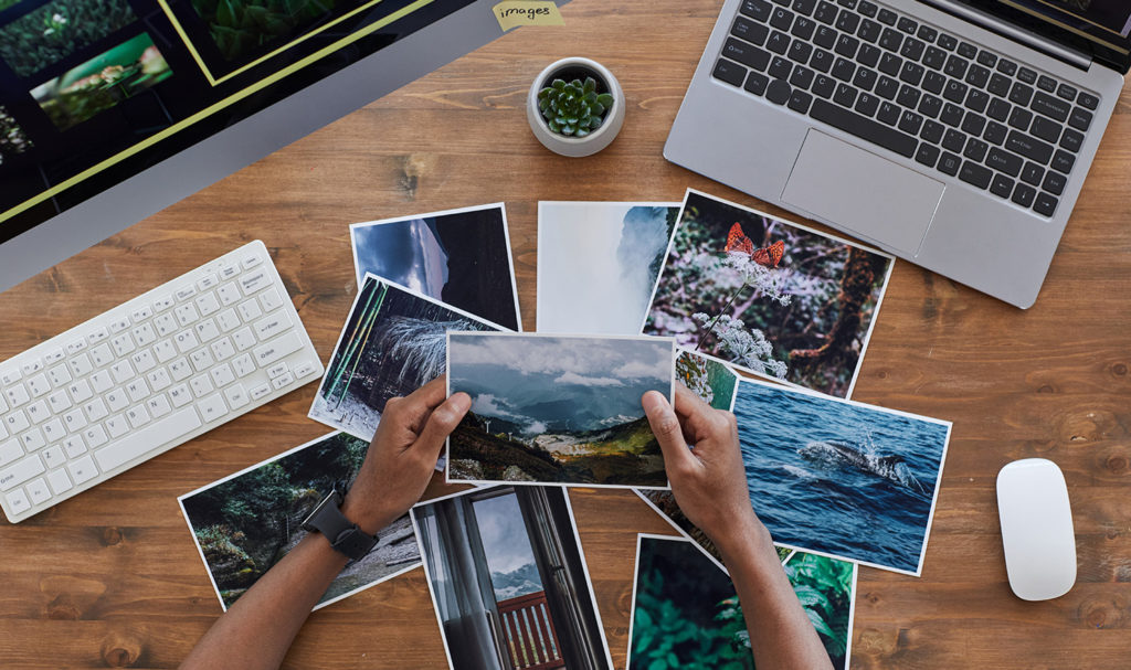 An overhead shot of a person holding a printed photo over a stack of prints laying on a wood desk next to a computer, coffee, and editing equipment. These prints can be insured with an ACT Annual policy.
