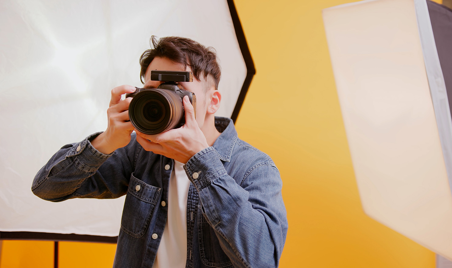 A waist up image of a young male photographer taking a photo with his camera aimed at you in a studio with a yellow backdrop and lighting equipment. ACT photography insurance may cover him at the next event he attends to sell his photos at.