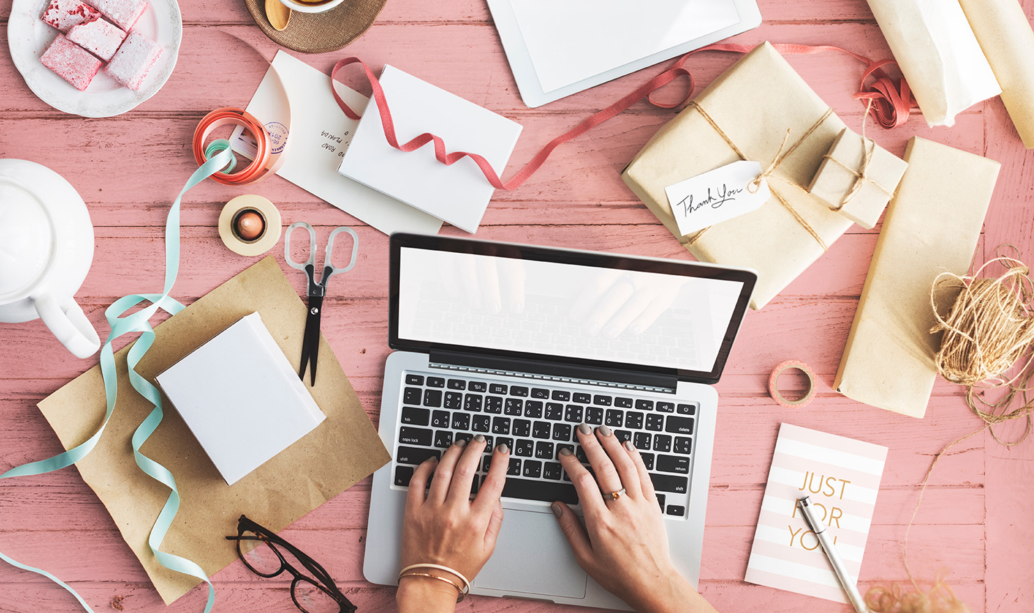 An overhead shot of a person working on a laptop on a pink wood table, with all their crafting supplies surrounding them.