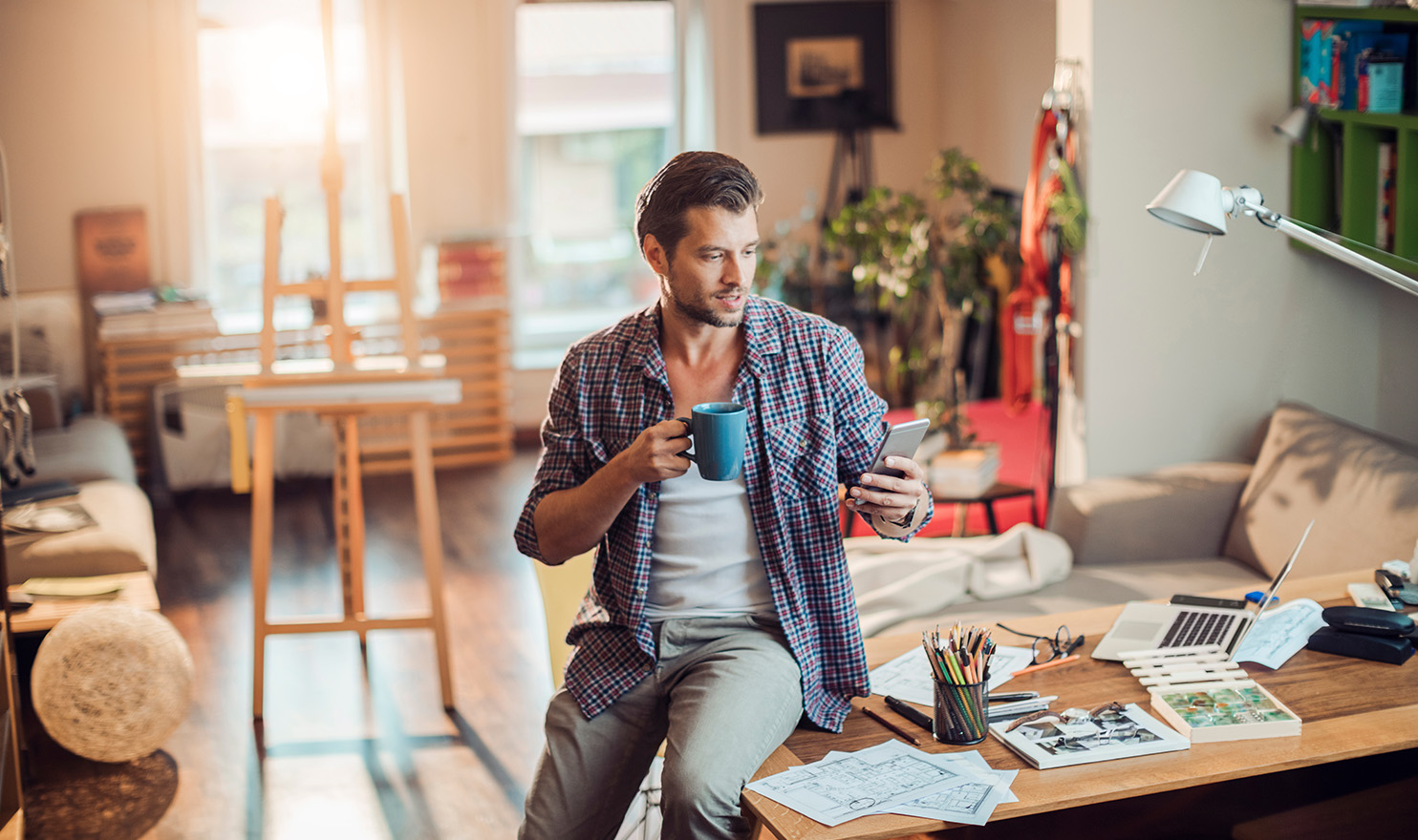 A crafter is sitting on the edge of a desk in his home studio where he sipping a mug of coffee and checking on his online craft sales on his cell phone.