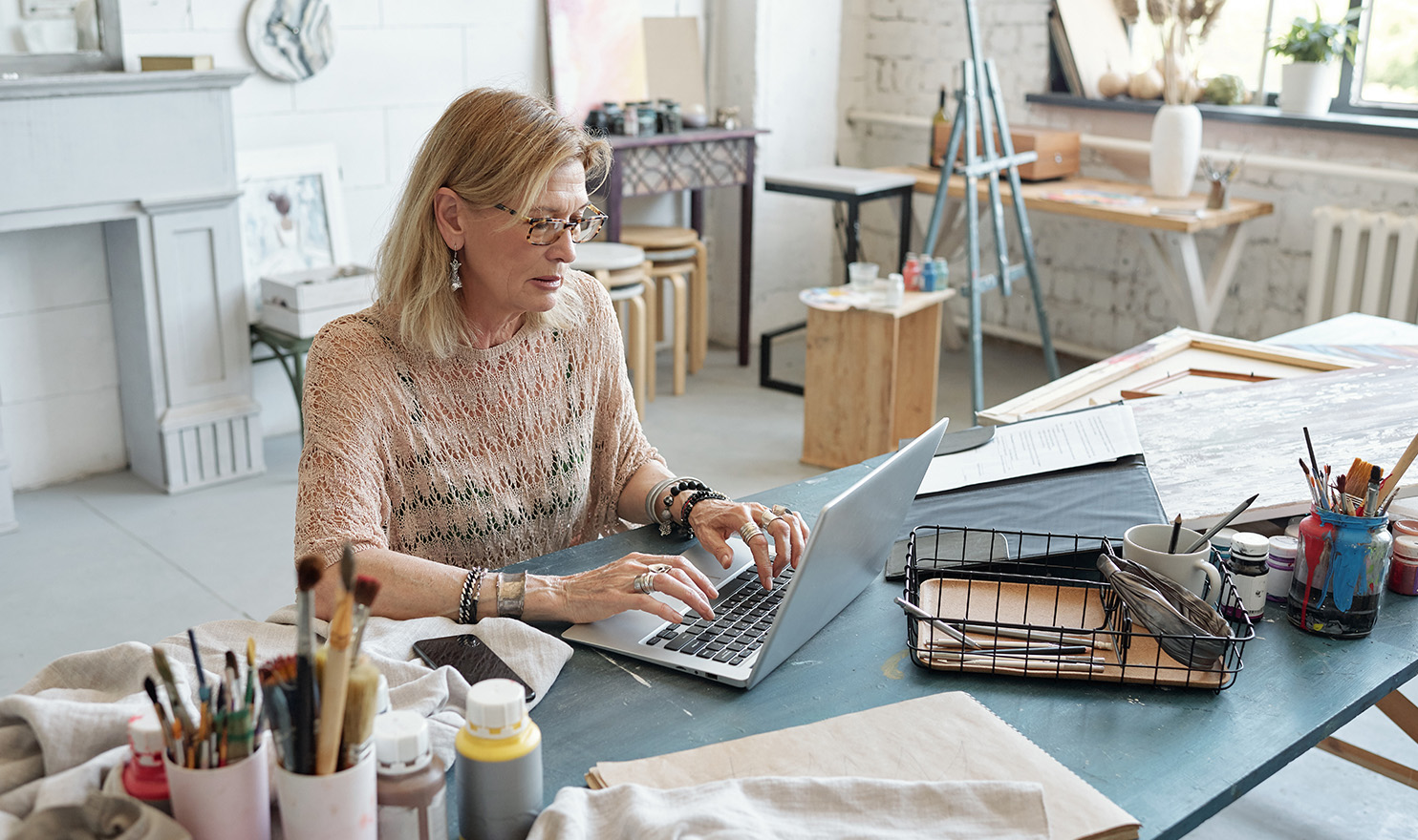 A crafter is working on a laptop in her home craft room, surrounding by her projects and tools.