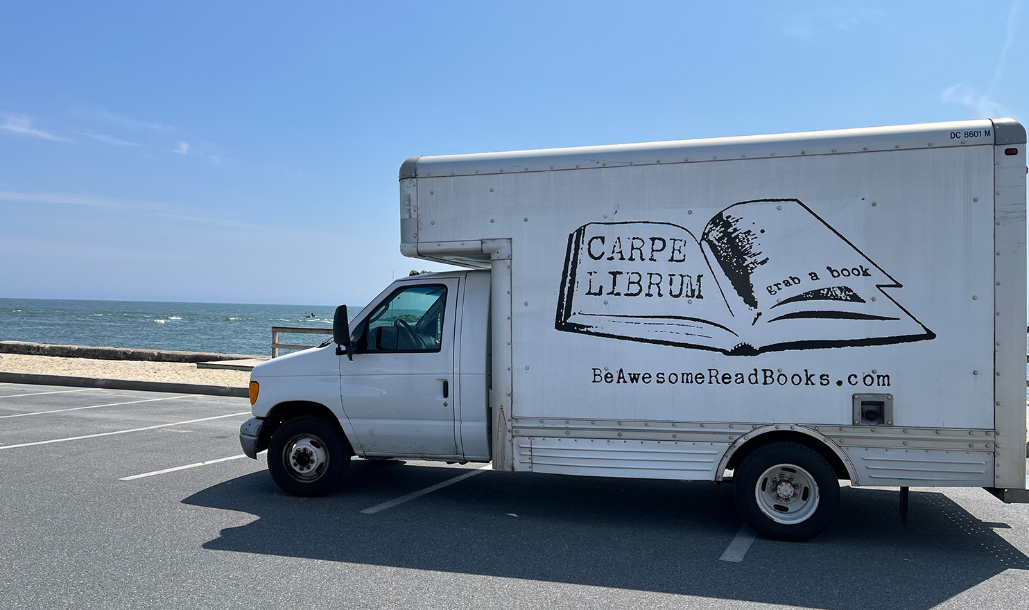 A white box truck with the Carpe Librum logo across the side is parked in a beach front parking lot. Elizabeth and her husband have invested in a truck to transport all their booth supplies to different states along the Eastern United States.
