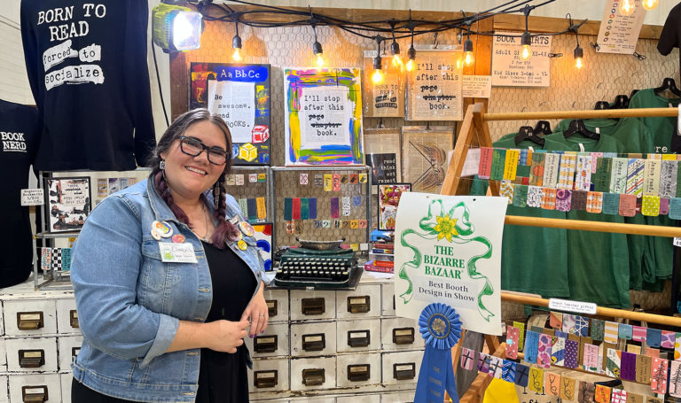 Elizabeth Fontenot standing in front of her booth for her business Carpe Librum at an event. Her display consists of vintage shelves, easels, and lights to display her handmade bookmarks and branded shirts and tote bags.