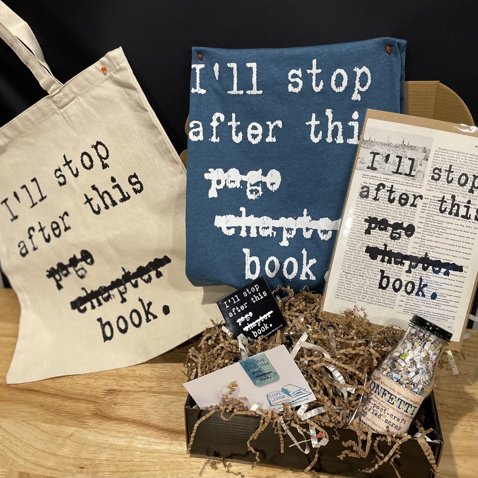 A display of Elizabeth's products include a tote bag, a tshirt, and a poster, all with the phrase, "I'll stop after this page, chapter, book," written on them. There are also bookmarks and a jar of recycled book confetti.