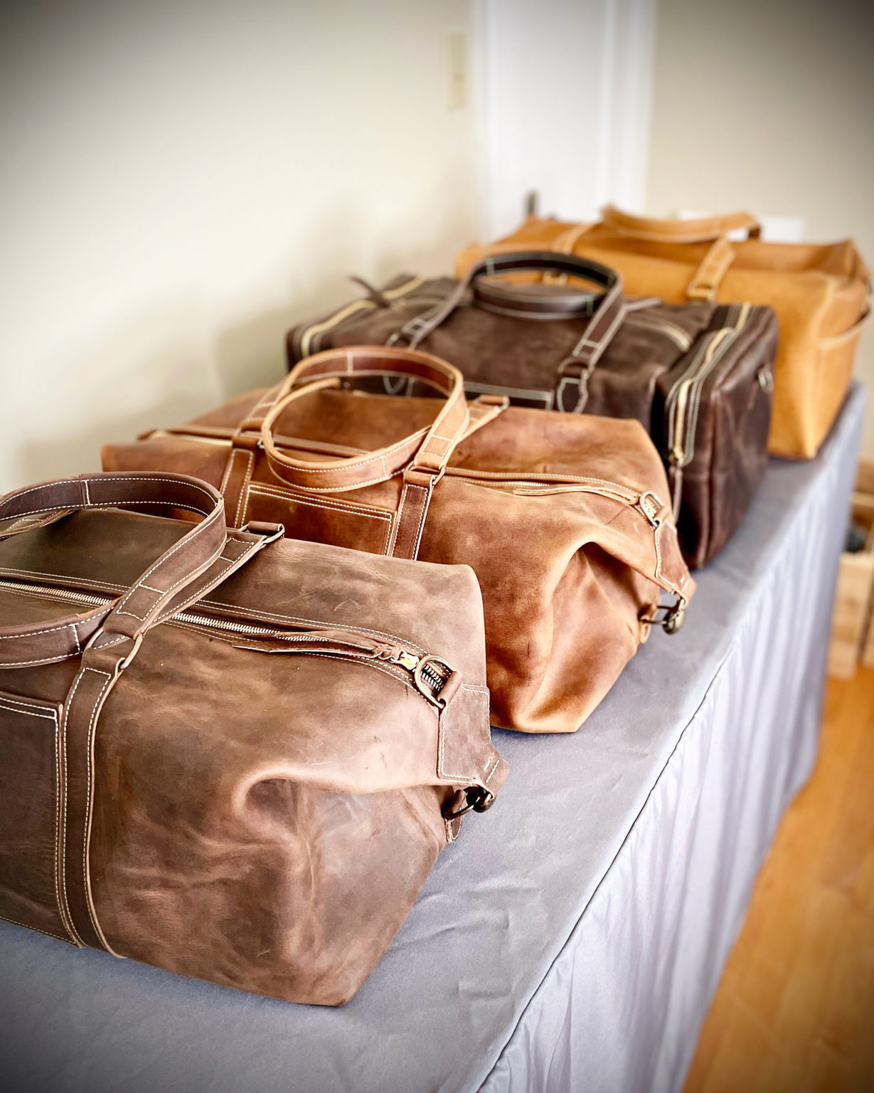 A variety of Kelsey's handcrafted leather bags, all made with a different type of leather, lay arranged in a row on a table at an event.