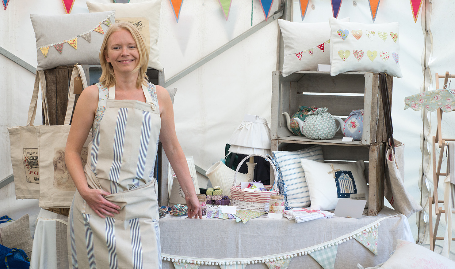A crafter stands proudly in her vendor booth at an event. She is wearing a striped apron and is standing in front of her display table of pillows, pottery, and home decor.