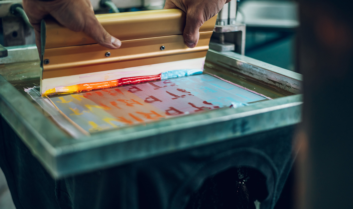 A close-up shot of an artist using teal, red, orange, and yellow ink to screen print a design onto a fabric object.
