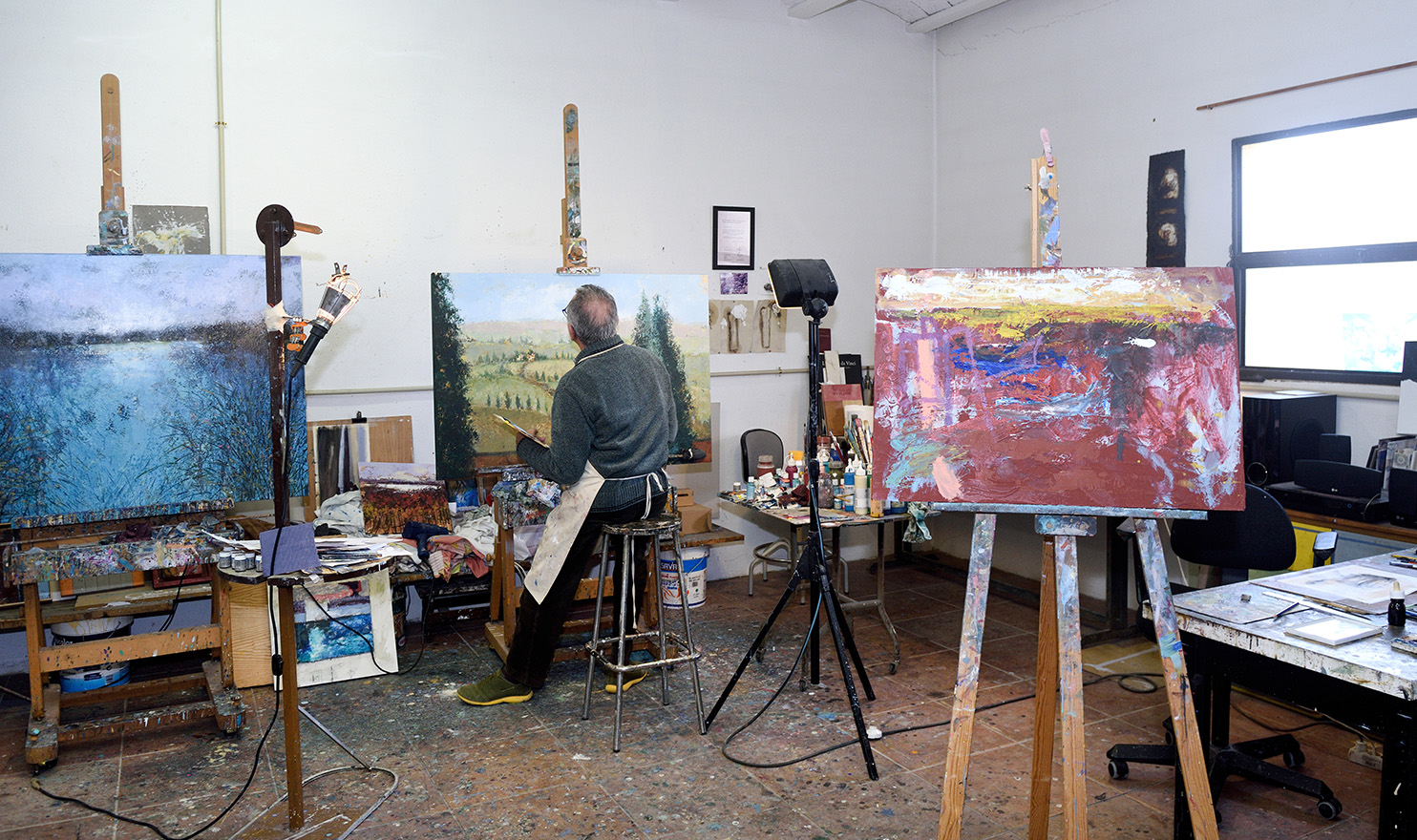 An artist has his back to the camera as he sits on a stool in an art studio and works on a landscape painting, with two empty easels beside him.