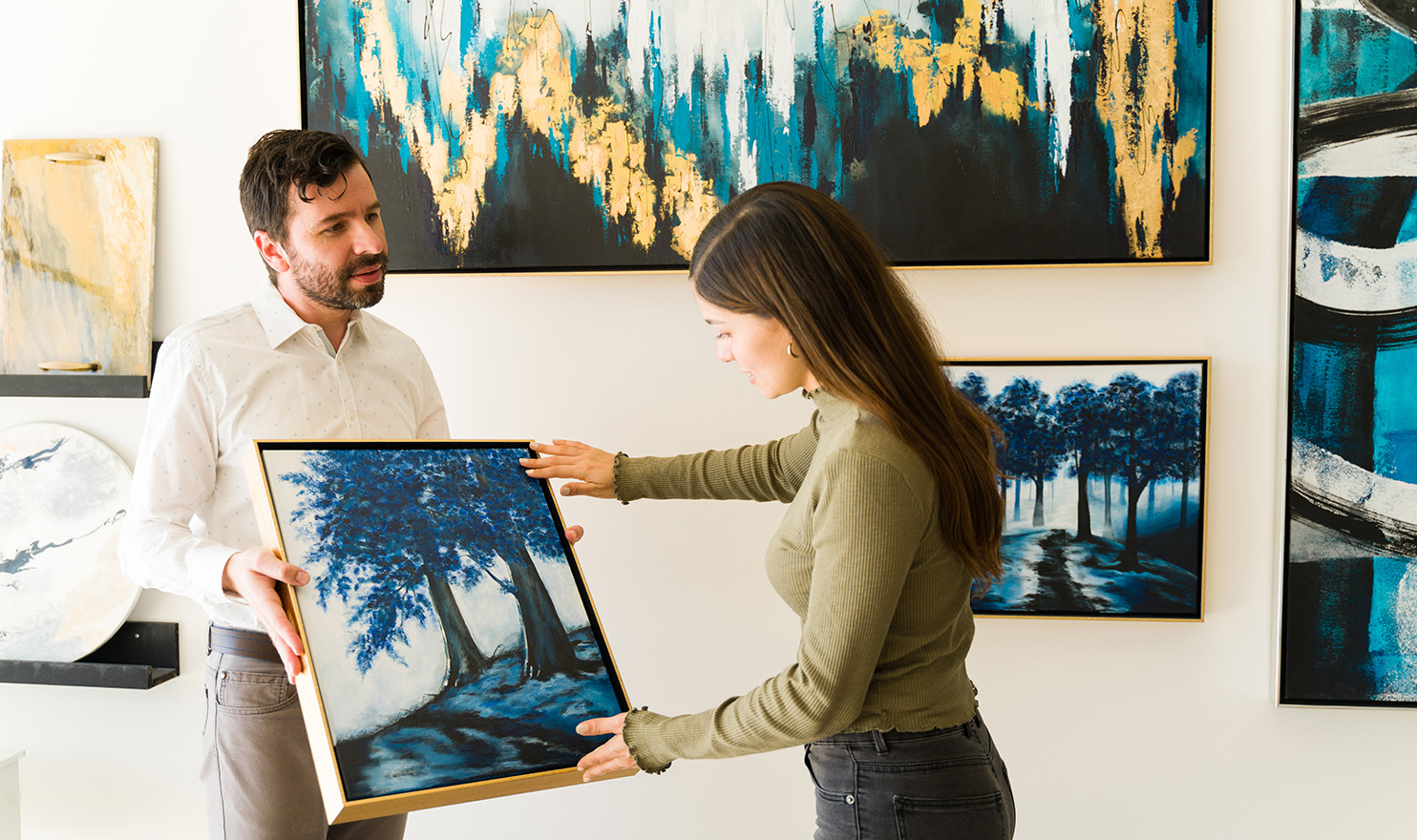 An artist is holding his painting that is part of a collection he is showcasing in a gallery. A potential buyer is examining his painting as she admires the detail in his work.