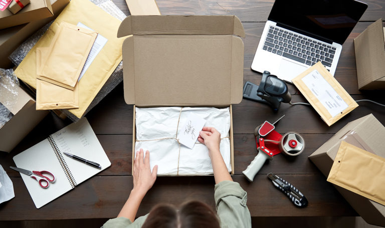 An overhead view of someone's hands packaging an order on their desk next to other shipping items and a laptop.