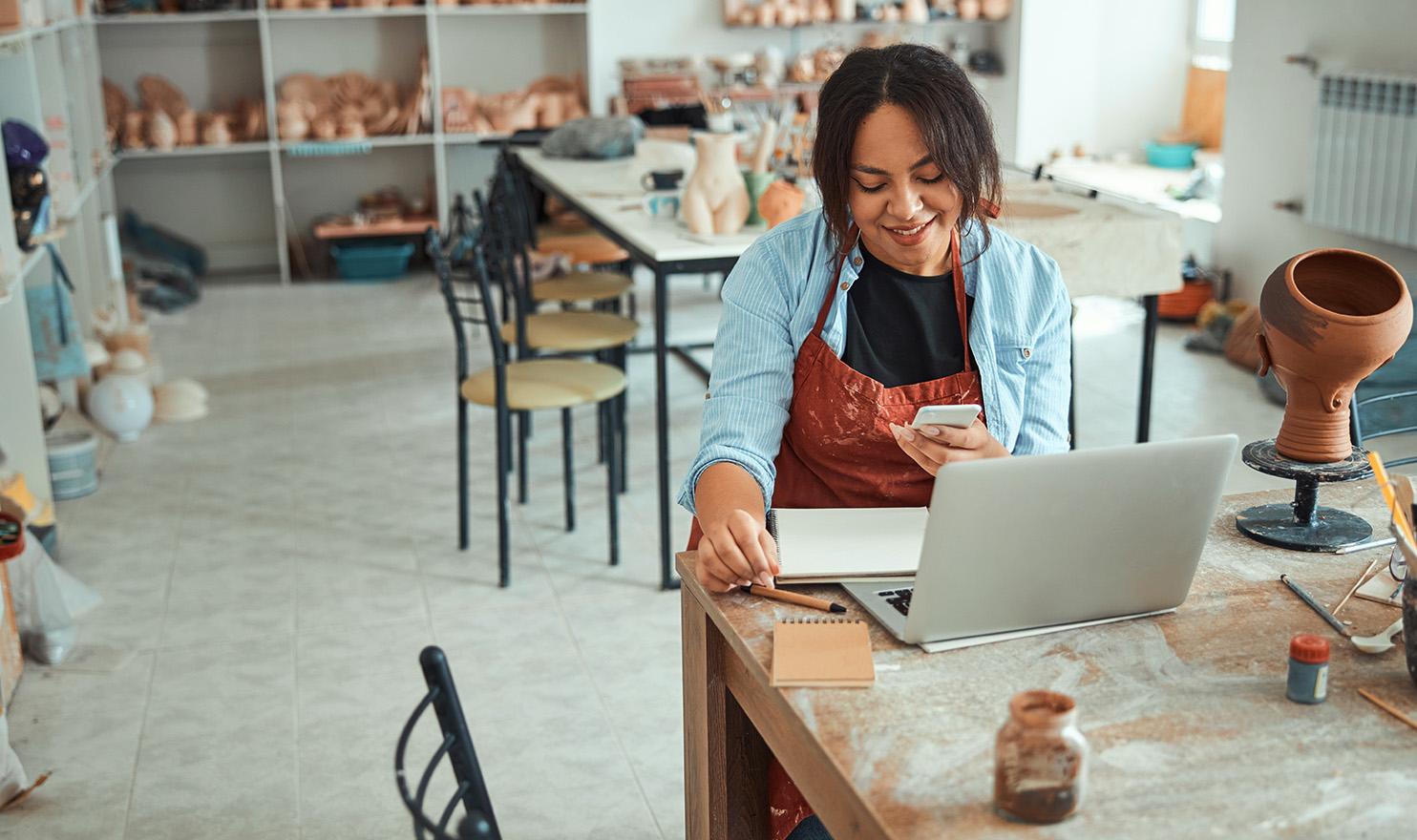 An artist is in her pottery studio happily working on her business from her smartphone and laptop.