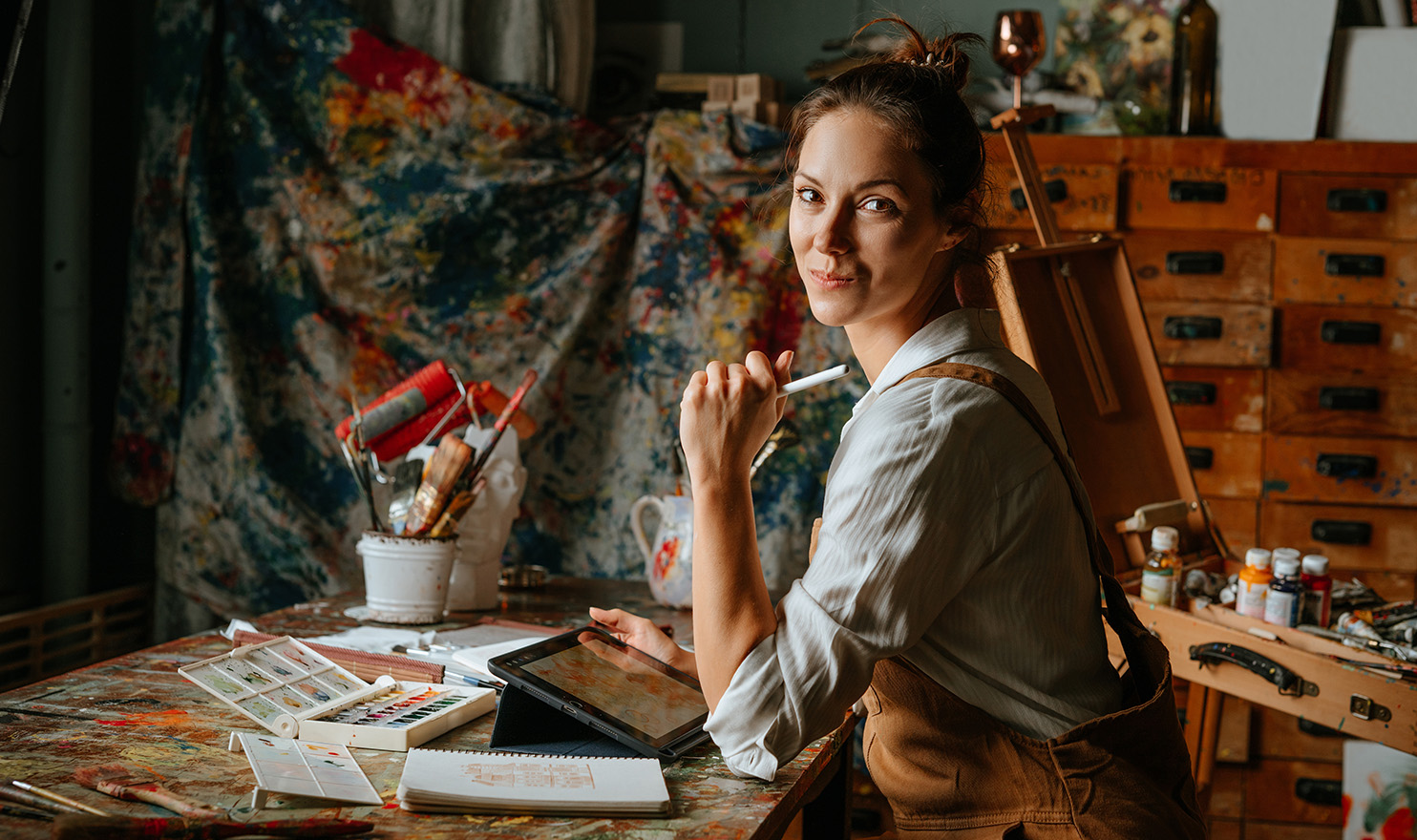 A artist is happily working on a laptop in her painting studio.