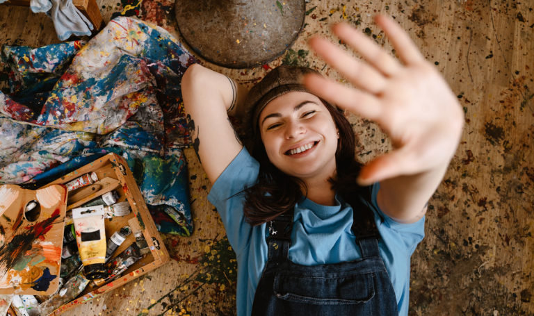 An artist is lying on the ground next to their art supplies while laughing with their hand in the air above them.