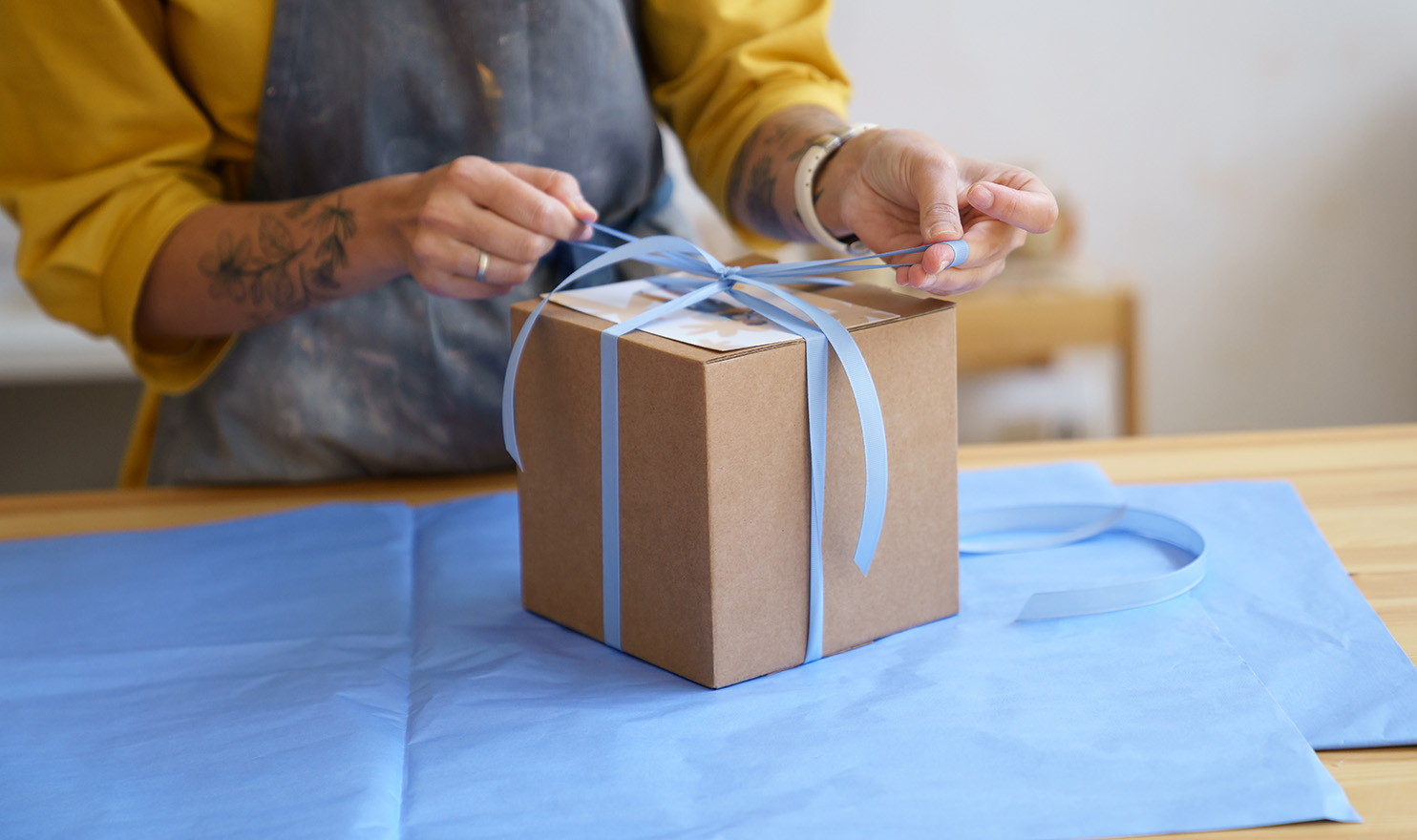 An artist is packaging an order and tying a blue ribbon around a box.