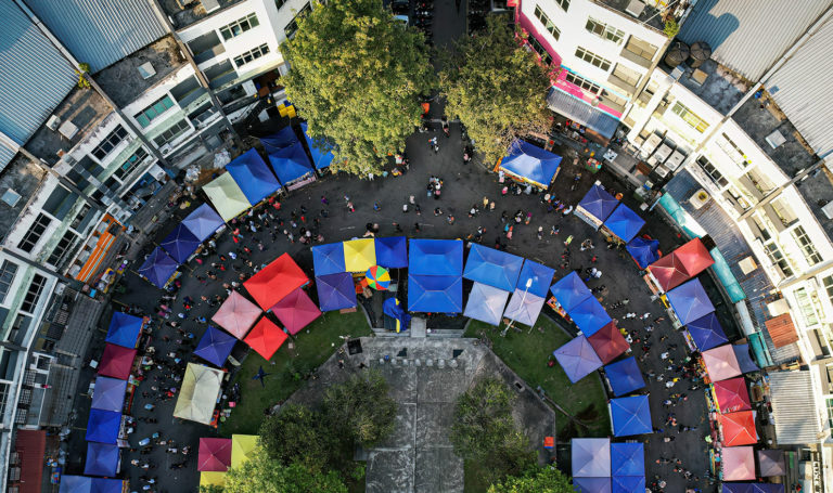 An overhead look at dozens of vendors lined up along a popular street in a city. They are selling their goods to customers underneath their colorful tents.