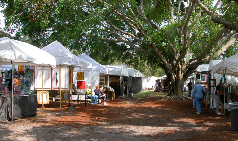 An outdoor arts and craft market with vendors and their white pop-up tents lining a shaded sidewalk.