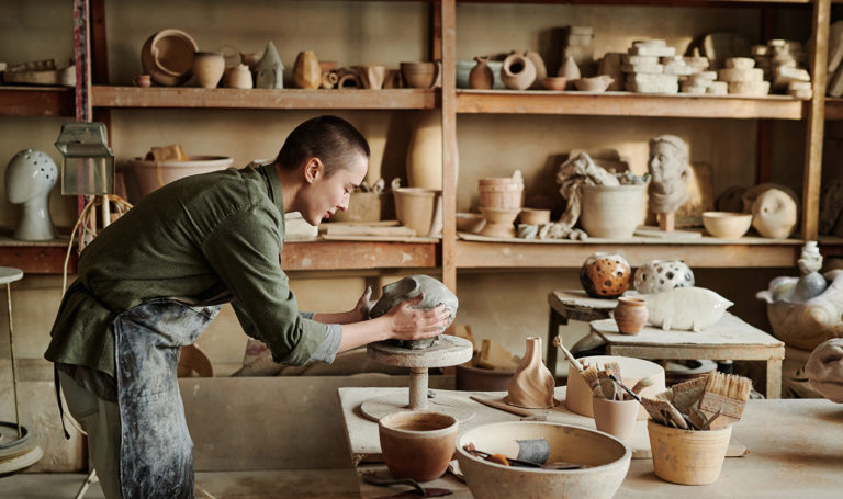 An artist is in his workshop leaning over a table as he focuses on the pottery piece he is making. He is surrounded by other pieces he has made that are on the table and on the shelves behind him.