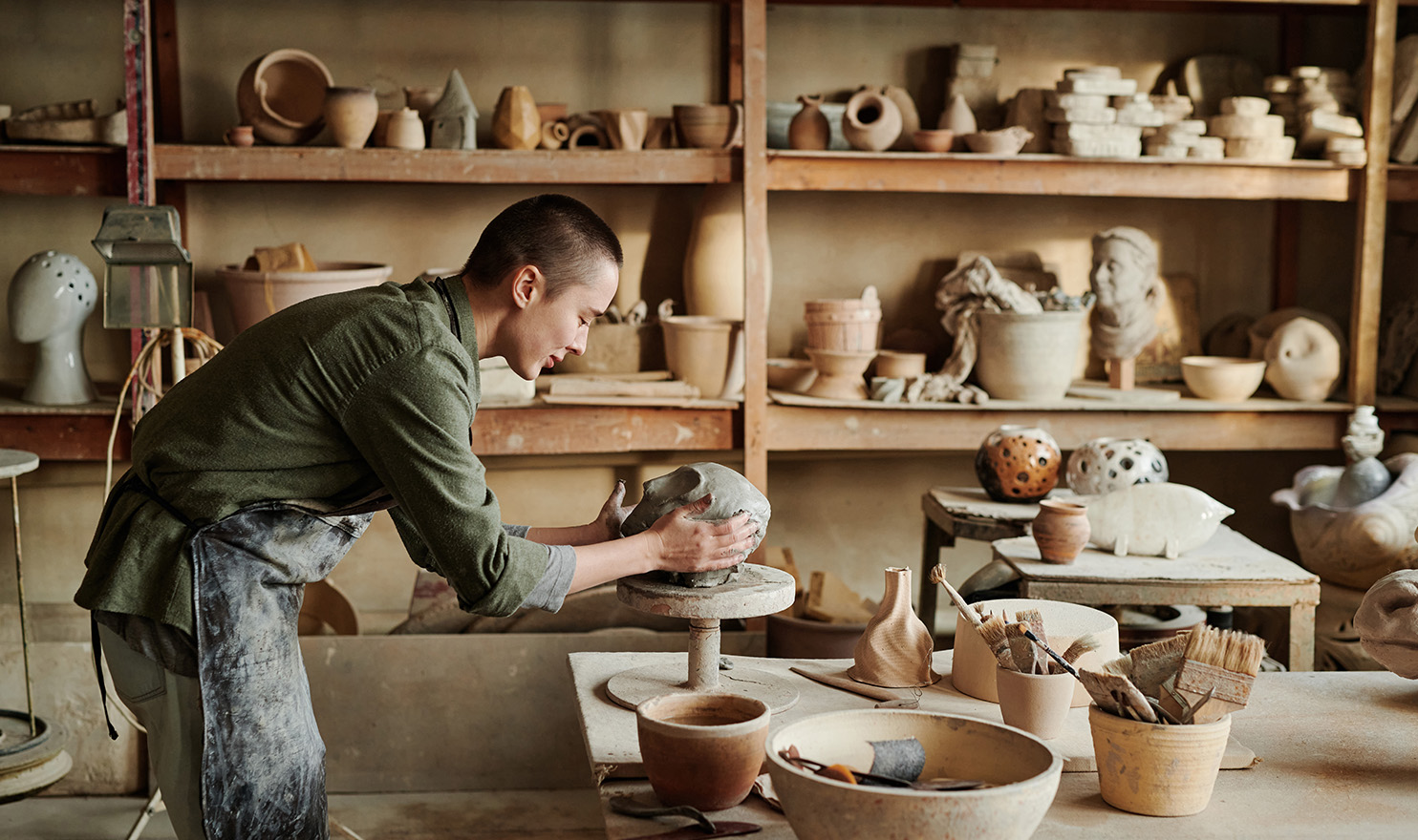An artist is in his workshop leaning over a table as he focuses on the pottery piece he is making. He is surrounded by other pieces he has made that are on the table and on the shelves behind him.