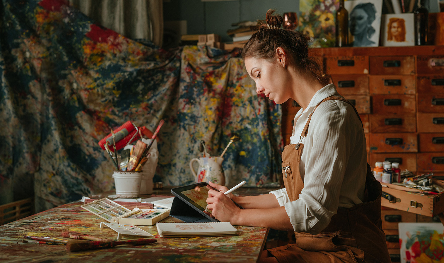 An artist is sitting at a table in her workshop as she completes business tasks on her tablet with a stylus pen. She is wearing her apron and is next to drop cloths she uses while painting.