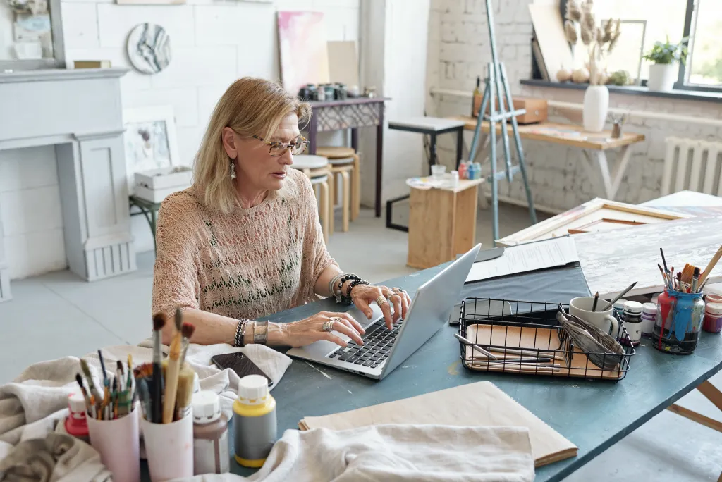 A female artist types at her laptop at a desk in her art studio. On the desk are cups of paintbrushes, canvas, and wooden frames. In the background, there are stools, an easel, potted plants, and a glass window.