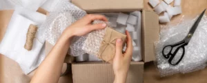 A crafter's hands inserting a small package wrapped in brown paper and twine into a bag of bubble wrap above a larger cardboard box with packing materials and scissors.