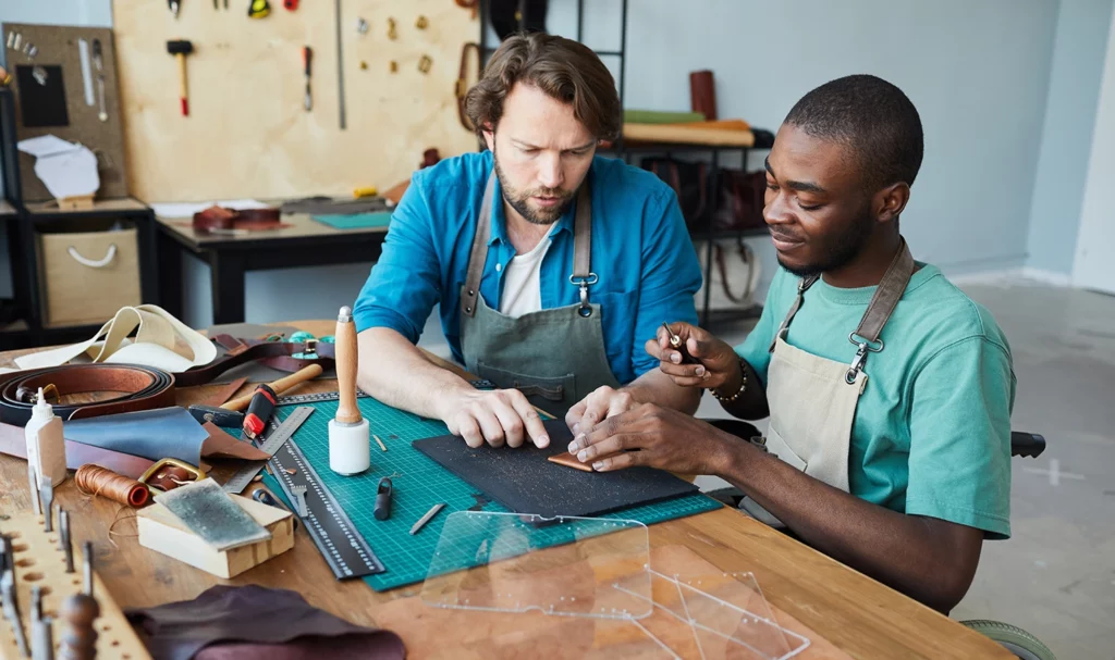 A leathercrafter carefully teaches an apprentice how to craft with certain leatherworking tools.