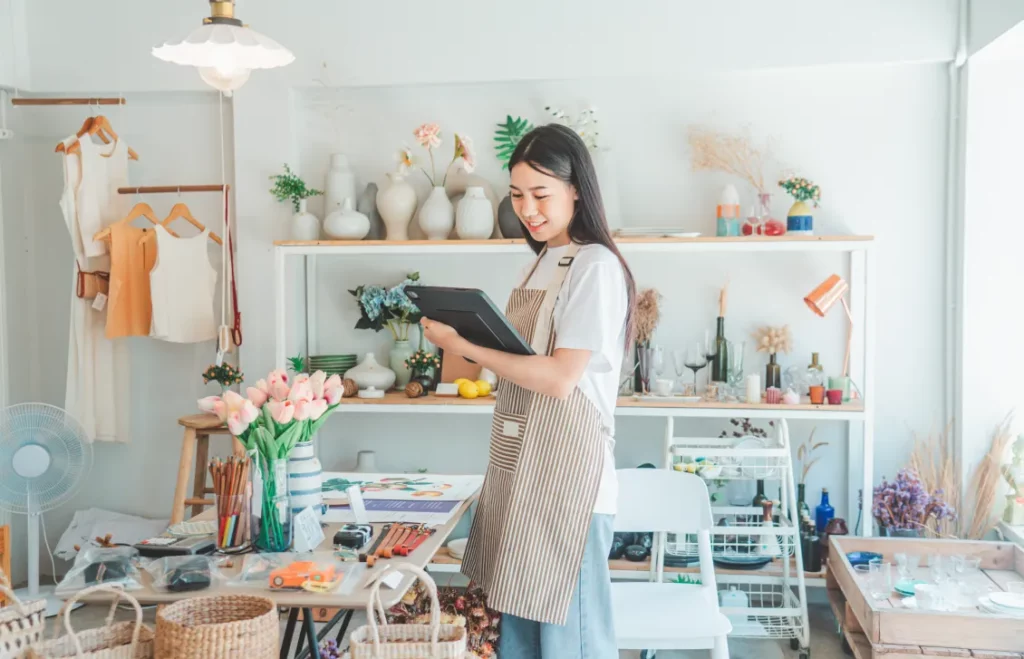 An artist wearing a white shirt, light denim jeans, and a brown stripe apron smiles while looking at a business tablet in the middle of an arts and crafts studio.