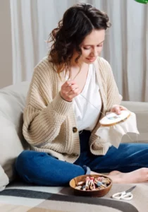 A woman wearing a cream cardigan and blue jeans threads an embroidery piece using a needle and embroidery hoop while sitting on a couch with supplies set in front of her.