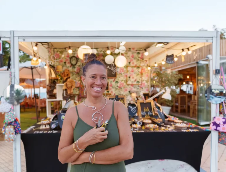 An artist wearing swirled metal jewelry and a sleeveless green dress holds pliers and smiles in front of her jewelry booth.