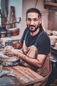 An artist wearing a black shirt and a brown apron works on a clay piece at a potter's wheel in a studio.