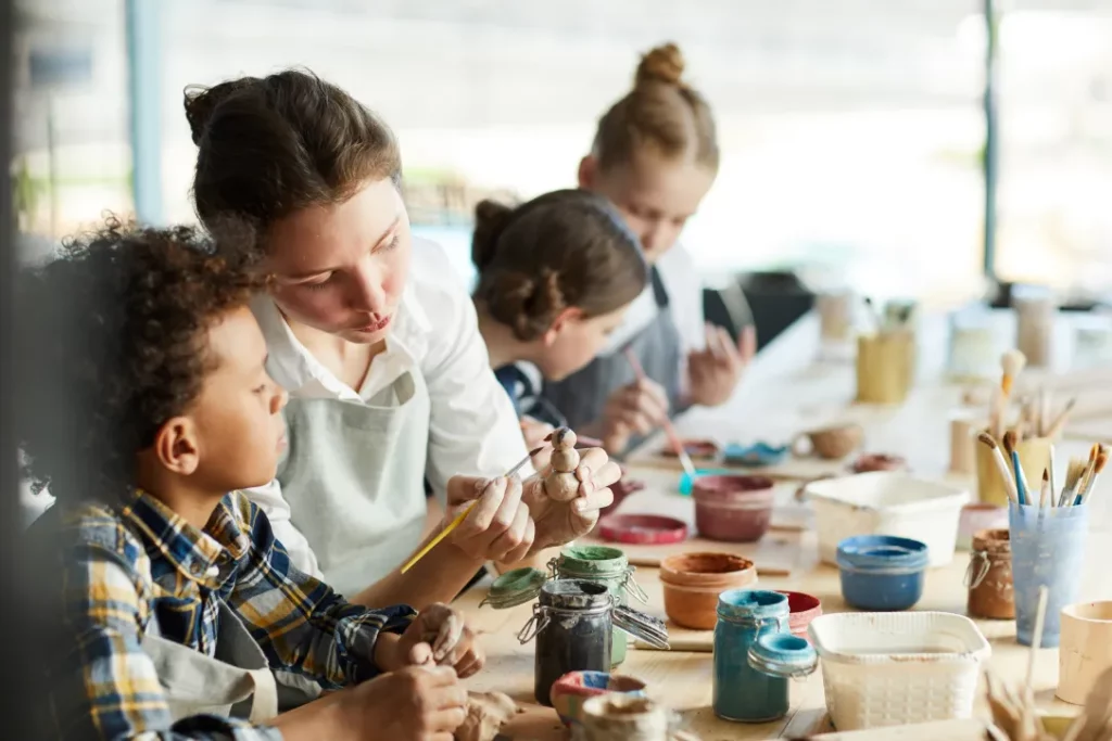 An artist shows a young workshop student how to paint a snowman sculpture at a table with various art supplies and other students.