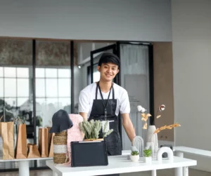 An artist wearing a black cap, a white shirt, and a black apron stands at his arts and crafts booth displaying ceramic vases and arches, a business tablet, plants, and raffia baskets.