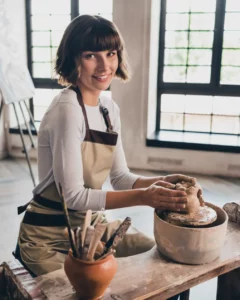 An artist wearing a white top and a beige apron shapes a piece of pottery in a studio.