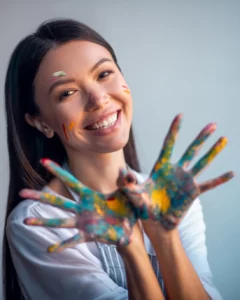 An artist smiles and shows the palms of her hands, which are covered in green, purple, and orange paint.