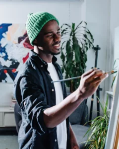 An artist wearing a green beanie and a denim shirt paints in a studio.