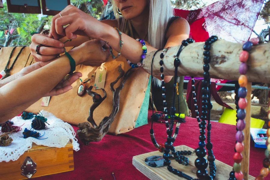 A crafts vendor clasps a bracelet on a customer's hand at a jewelry booth displaying jewelry on wooden stands on a hot pink cloth table.