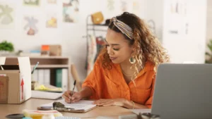 young female business owner sitting at a laptop while writing on a piece of paper on a clipboard