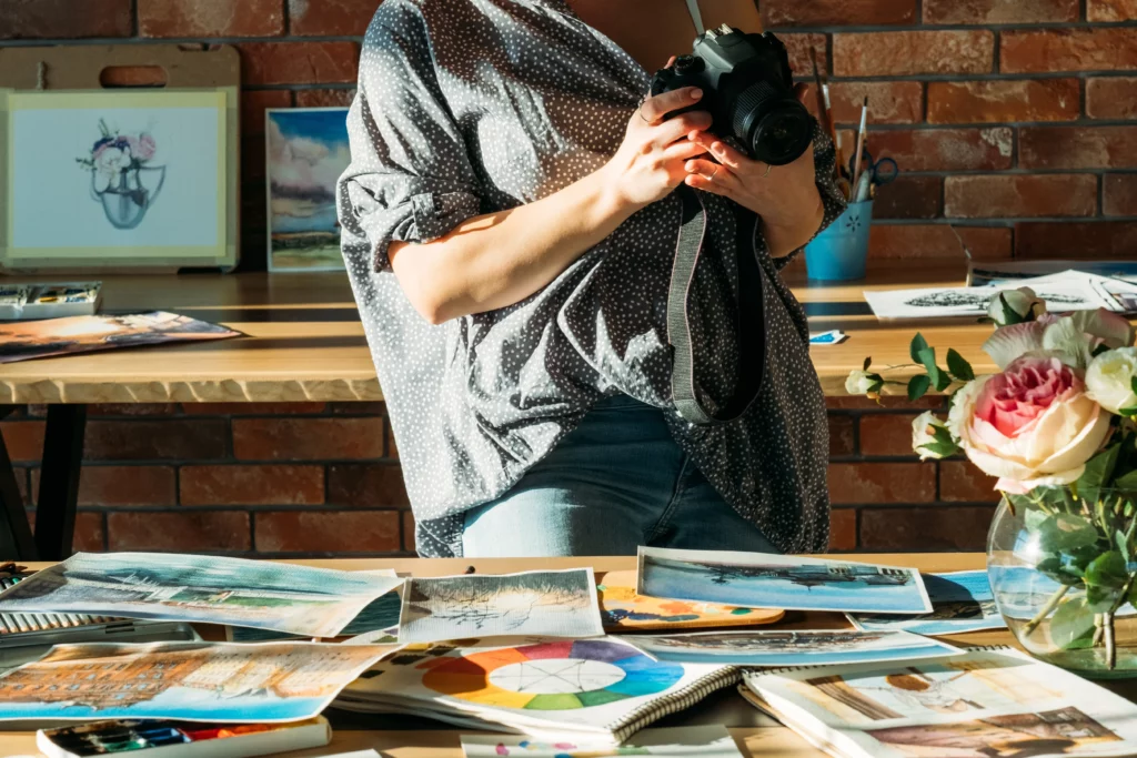 An artist wearing a polka dot blouse and blue jeans holds a camera above a workspace desk with various art prints, notebooks, and a pink flower bouquet in an art studio.