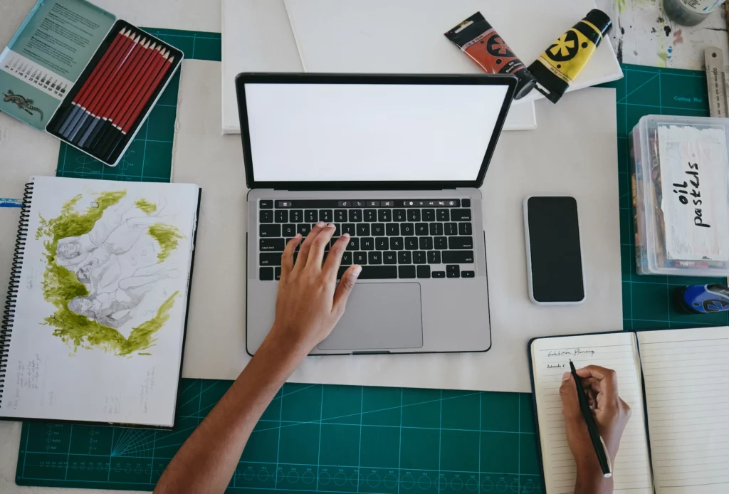 An overhead shot of an artist typing on a laptop and writing in a notebook on a desk with a tin of red pencils, paint tubes, a smartphone, a green grid desk pad, and a notebook with an art sketch colored with green paint.