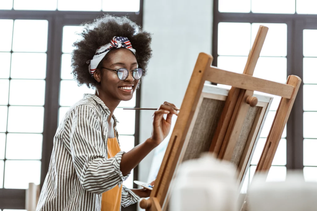 An artist wearing a patterned headscarf, a striped shirt, and an orange blouse smiles while painting on a canvas in a studio with large windows in the background.