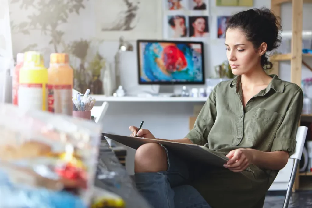 An artist sits and writes notes on a large writing pad in an art studio with various art supplies set on a table in the foreground.