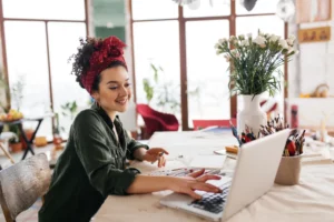 An artist wearing a green shirt and red polka dot bandana smiles and taps on her laptop while sketching with markers at her studio desk.