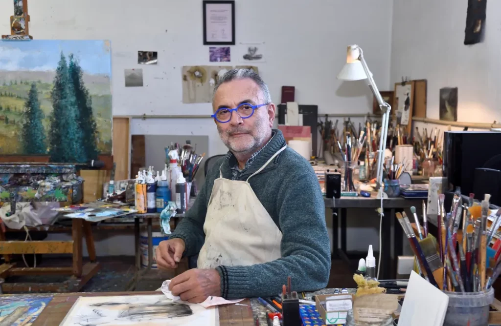 An artist wearing a green sweater and beige apron sits at a wooden desk in his art studio with a canvas depicting green trees, plus various paintbrushes and art tools in the background.