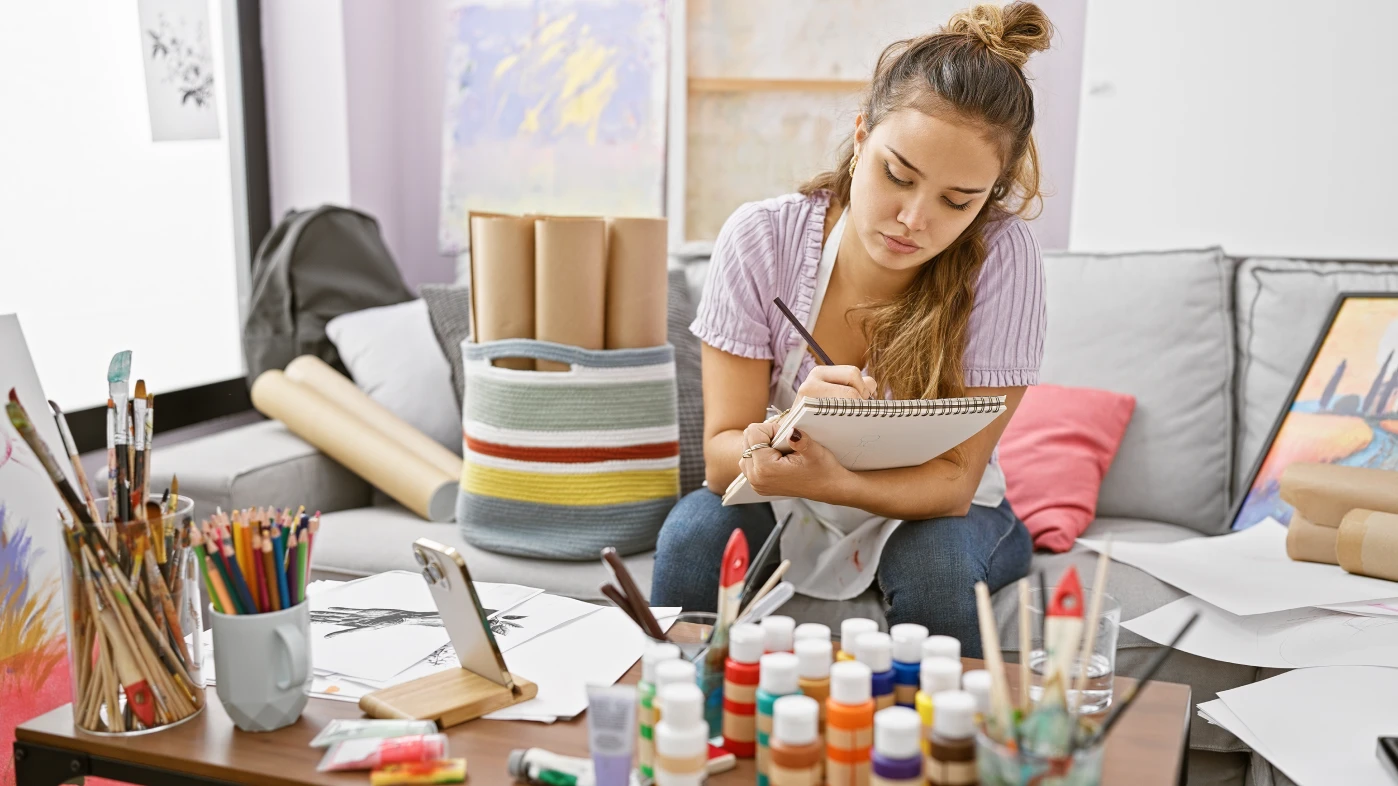 a young female artist working in a sketchbook while surrounded by art supplies